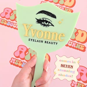 Puede incluir: Un espejo de mano verde claro con el nombre "Yvonne" en letras doradas y las palabras "Eyelash Beauty" debajo. El espejo presenta un diseño de ojo y dos estrellas doradas. También se ve el texto "comes in SEVEN stunning shades!"