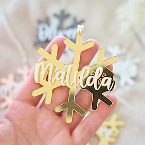 May include: A gold-coloured snowflake ornament with the name "Matilda" in white script. The ornament is held in a hand, with other snowflake ornaments visible in the background. A small string is attached for hanging.
