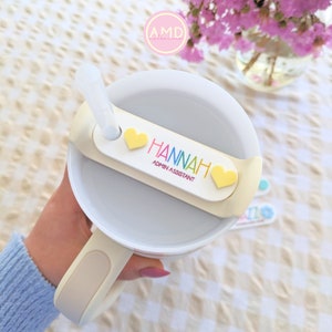 May include: A white travel mug with a beige handle and a straw. The mug has a yellow name tag with the text "HANNAH ADMIN ASSISTANT" and two yellow hearts.
