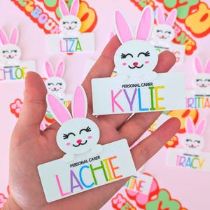 May include: White bunny rabbit name tags with colourful text. The name tags say "Kylie", "Lachie", "Chloe", "Liza", "Tracy", "Britt", and "Christine". Each name tag has the text "Personal Carer" above the name.