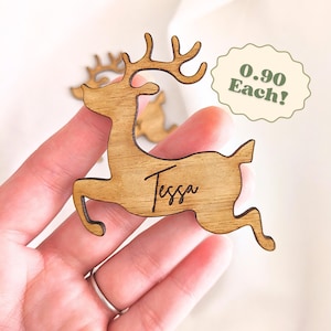 May include: A wooden reindeer cutout with the name "Tessa" engraved on its side. The reindeer is light brown and has detailed antlers. The image shows the reindeer being held by a person's hand.