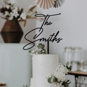 May include: A black cake topper reading "The Smiths" in cursive script, placed on a white tiered cake adorned with white roses and foliage. The cake is the central element, decorated for a celebration.