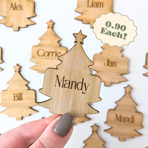 Può includere: Etichette con nome a forma di albero di Natale in legno con nomi incisi. Il nome "Mandy" è ben visibile su un'etichetta. Altre etichette includono nomi come "Alex", "Liam" e "Corrie". Ogni etichetta ha una stella in cima. È visibile anche il testo "0.90 Each!"