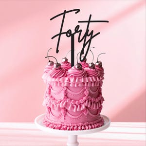 以下が含まれることがあります： ピンク色のケーキ。デコレーションされたフロスティングと黒い「Forty」のケーキトッパーが付いています。ケーキは銀色のチェリーで飾られています。ピンク色の背景に白いケーキスタンドに載っています。お祝いのデザートです。