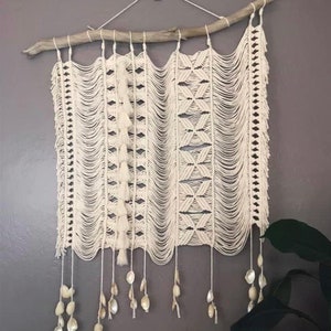 May include: Cream-colored macrame wall hanging. The artwork is suspended from a natural wooden branch. The design features intricate knotting patterns, tassels, and dangling shell accents, creating a bohemian aesthetic.