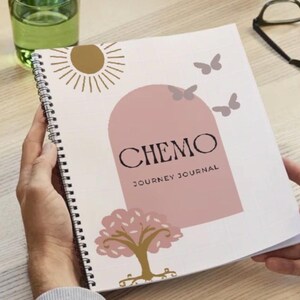 May include: A spiral-bound journal with a cover featuring the text "CHEMO JOURNEY JOURNAL" in black. The design includes a pink arch, butterflies, a sun, and a tree. The journal is held open by hands.