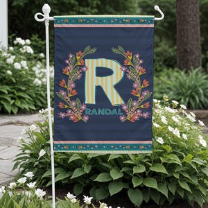 May include: A navy blue garden flag featuring a floral wreath design. The letter "R" is in the centre, with yellow and white stripes. The name "Randal" is below the letter. The flag is hanging on a white metal stand.
