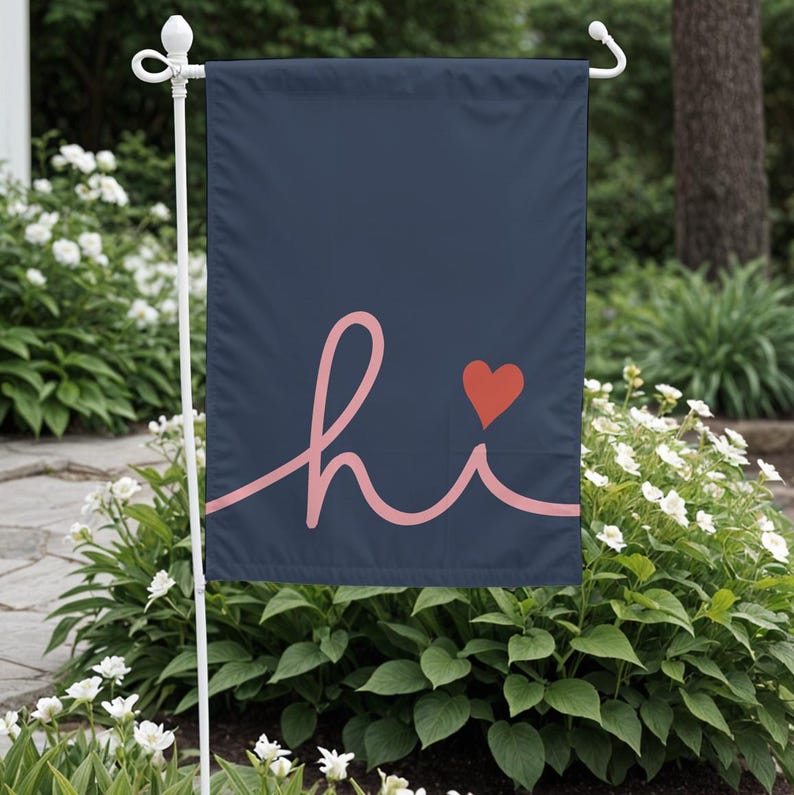 Pode incluir: Uma bandeira de jardim azul marinho com a palavra "hi" em it&aacute;lico coral e um pequeno cora&ccedil;&atilde;o vermelho. A bandeira &eacute; exibida em um suporte de metal branco, cercada por plantas verdes e flores brancas.