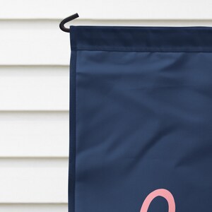 Pode incluir: Uma bandeira de jardim azul marinho com uma letra cursiva rosa. A bandeira est&aacute; pendurada em um gancho de metal preto. O fundo &eacute; um revestimento horizontal branco.