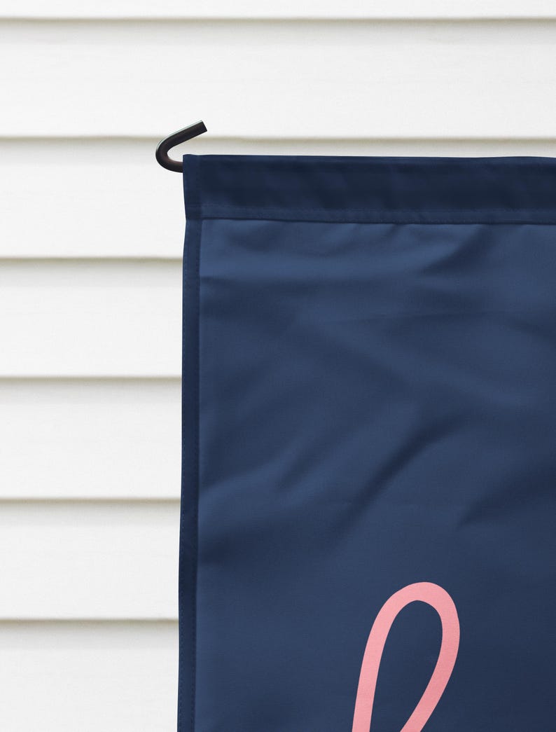 Pode incluir: Uma bandeira de jardim azul marinho com uma letra cursiva rosa. A bandeira est&aacute; pendurada em um gancho de metal preto. O fundo &eacute; um revestimento horizontal branco.