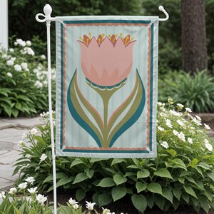 Peut inclure: Un drapeau de jardin avec un motif de tulipe rose et des accents turquoise et verts. Le drapeau a un fond &agrave; rayures bleu clair et une bordure festonn&eacute;e. Il est affich&eacute; sur un support en m&eacute;tal blanc, entour&eacute; de verdure et de fleurs blanches.