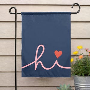 Pode incluir: Uma bandeira de jardim azul marinho com a palavra "hi" em rosa claro em it&aacute;lico e um pequeno cora&ccedil;&atilde;o vermelho. A bandeira est&aacute; pendurada em um suporte de metal preto, com um fundo de revestimento de madeira.