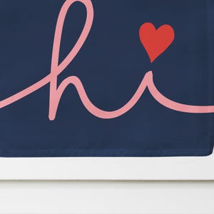 Pode incluir: Uma bandeira de jardim azul marinho com a palavra "hi" em fonte cursiva rosa claro. Um cora&ccedil;&atilde;o vermelho est&aacute; acima do "i". A bandeira est&aacute; pendurada em um poste de metal preto contra um fundo branco.