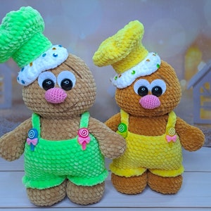 May include: Two crocheted plush toys shaped like gingerbread people. The toy on the left is brown with green overalls and a green chef's hat. The toy on the right is brown with yellow overalls and a yellow chef's hat.
