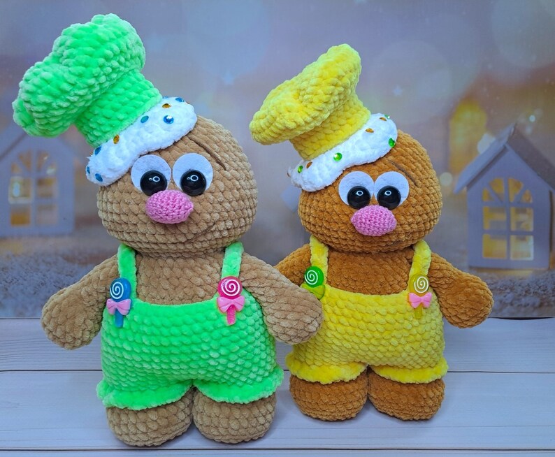 May include: Two crocheted plush toys shaped like gingerbread people. The toy on the left is brown with green overalls and a green chef's hat. The toy on the right is brown with yellow overalls and a yellow chef's hat.