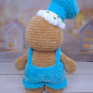 May include: A crocheted brown teddy bear wearing a blue hat and blue overalls. The hat has white frosting and colorful sprinkles.