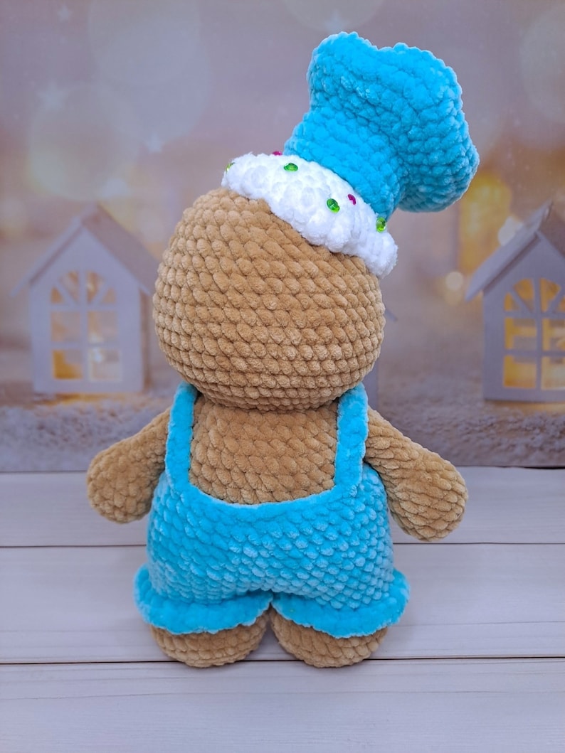 May include: A crocheted brown teddy bear wearing a blue hat and blue overalls. The hat has white frosting and colorful sprinkles.