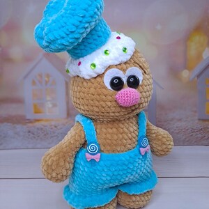 May include: A crocheted gingerbread man toy wearing a blue chef's hat, blue overalls, and a pink nose. The toy has large black eyes and a white face with colorful sprinkles.