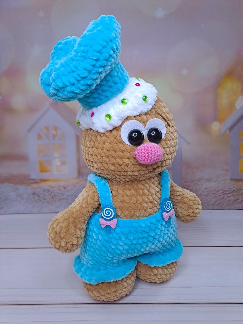 May include: A crocheted gingerbread man toy wearing a blue chef's hat, blue overalls, and a pink nose. The toy has large black eyes and a white face with colorful sprinkles.