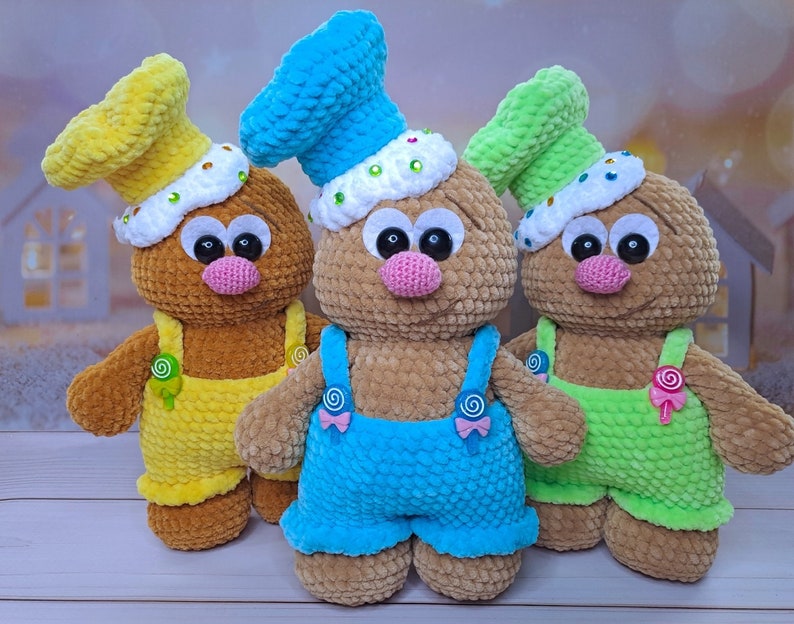 May include: Three crocheted plush toys shaped like gingerbread people. The toys are wearing different colored overalls and hats. The toys are brown, yellow, and green.
