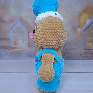 May include: A crocheted gingerbread person toy with a blue hat and overalls. The toy has a pink nose and brown fur.