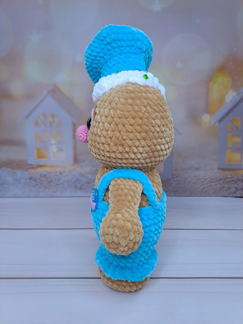 May include: A crocheted gingerbread person toy with a blue hat and overalls. The toy has a pink nose and brown fur.