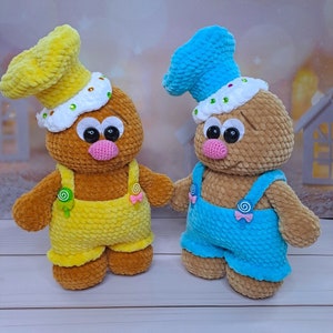 May include: Two crocheted gingerbread people toys with brown bodies, yellow and blue overalls, and white frosting hats. The gingerbread person on the left has a yellow hat and yellow overalls, while the gingerbread person on the right has a blue hat and blue overalls.