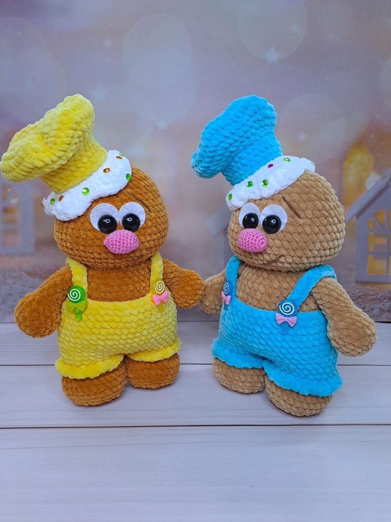 May include: Two crocheted gingerbread people toys with brown bodies, yellow and blue overalls, and white frosting hats. The gingerbread person on the left has a yellow hat and yellow overalls, while the gingerbread person on the right has a blue hat and blue overalls.