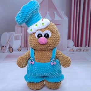 May include: A crocheted plush toy with a brown body, blue overalls, and a blue and white chef's hat. The toy has large, black eyes and a pink nose.