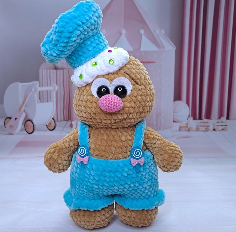 May include: A crocheted plush toy with a brown body, blue overalls, and a blue and white chef's hat. The toy has large, black eyes and a pink nose.