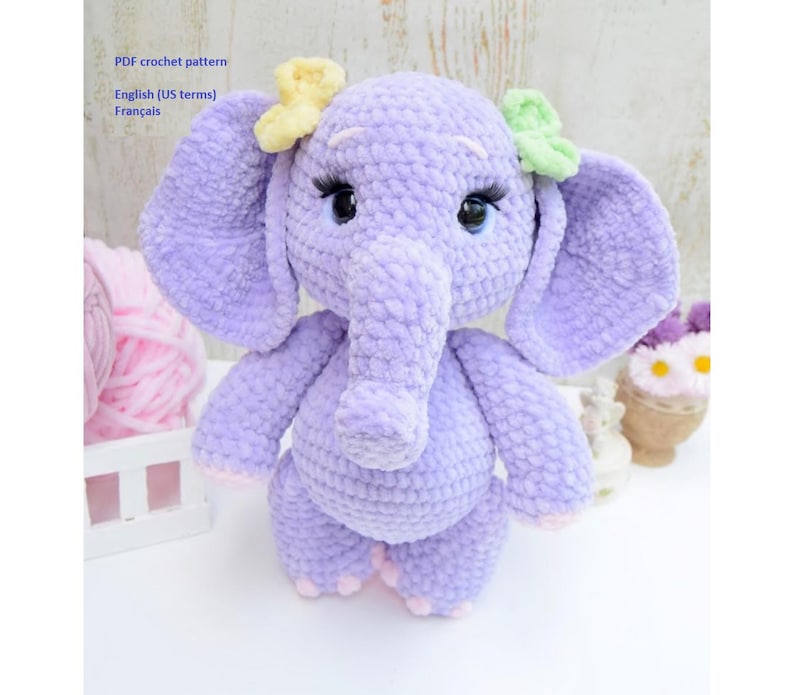 May include: A crocheted purple elephant toy with a long trunk and big ears. The elephant has pink feet and is decorated with yellow and green flowers.