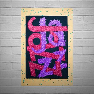 May include: A framed artwork with a black background and the word "JAZZ" spelled out in pink and purple fuzzy letters. The frame is made of wood and has a light brown colour. The frame is decorated with small turquoise and yellow confetti.