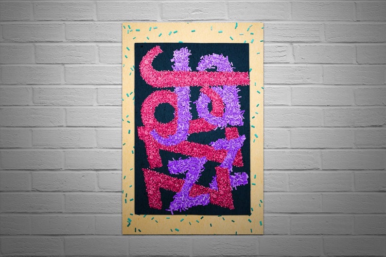 May include: A framed artwork with a black background and the word "JAZZ" spelled out in pink and purple fuzzy letters. The frame is made of wood and has a light brown colour. The frame is decorated with small turquoise and yellow confetti.