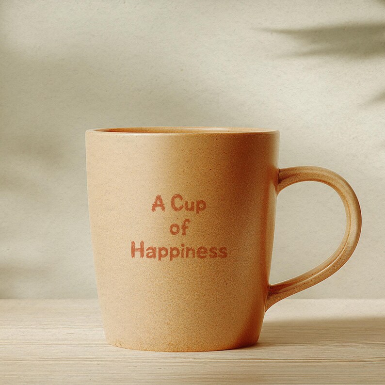 May include: A beige ceramic mug with a curved handle. The mug features the text "A Cup of Happiness" in a matching beige colour. The mug is set against a neutral background, suggesting a cosy, inviting atmosphere.