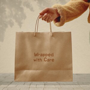 May include: A brown paper shopping bag with twisted paper handles is held. The bag has the words "Wrapped with Care" printed on the front. The person is wearing a tan jacket.