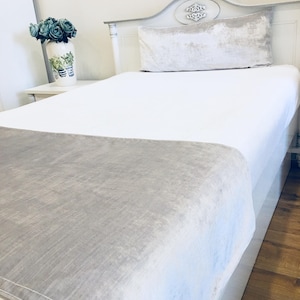 May include: A white twin-sized bed with a gray velvet headboard and a gray velvet bolster pillow. The bed is made with white sheets and a gray velvet throw blanket.