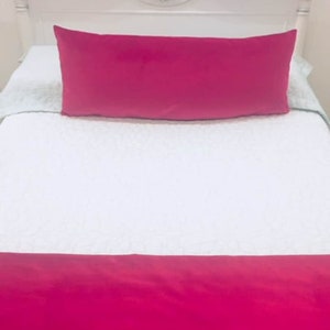 May include: A white bed with a pink pillow and a pink bed skirt. The headboard is white with a decorative design.