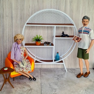 May include: A dollhouse scene featuring a white circular bookshelf with brown shelves, a doll reading a book in an orange chair, and a doll holding a book. The bookshelf is decorated with a plant, books, and a blue bottle. The dolls are dressed in casual attire.