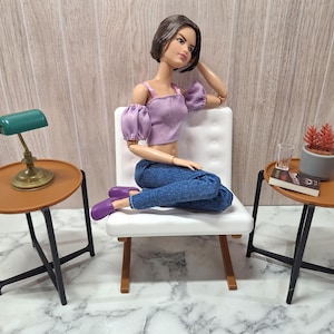 Puede incluir: Una muñeca con un top lila y vaqueros azules está sentada en una silla blanca. Dos pequeñas mesas marrones con patas negras flanquean la silla. Una tiene una lámpara verde, la otra una planta y un libro.