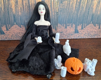 May include: A doll in a black dress with long black hair, holding a white candle. Several white candles, a carved orange pumpkin, and a white skull are arranged around the doll. The background features a forest scene.