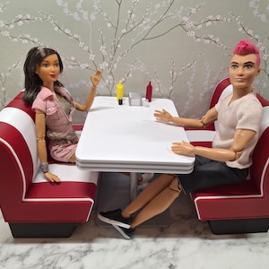 May include: A miniature diner scene with two dolls seated at a white table. The booth seating is red and white. The dolls are dressed in casual clothes. Condiments are on the table. The background is a floral wallpaper.
