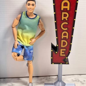 Könnte beinhalten: Eine Puppe in einem blau-gelben Tanktop und Jeansshorts, die an einem rot-schwarzen Arcade-Schild lehnt. Das Schild trägt das Wort "ARCADE" in gelben Buchstaben und ist mit gelben Lichtern verziert.