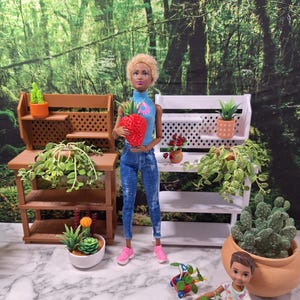 Puede incluir: Una muñeca con una blusa azul y vaqueros sostiene una gran fresa roja. Pequeños soportes de madera para plantas y macetas con plantas están dispuestos alrededor de la muñeca. Una segunda muñeca está sentada, jugando con un vagón de juguete lleno de plantas. El fondo es una escena de bosque.