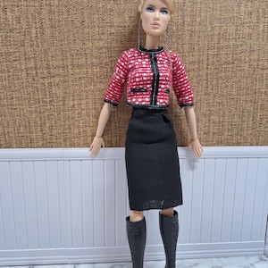 Puede incluir: Una muñeca vestida con una chaqueta estampada en rojo y blanco con ribetes negros, una falda lápiz negra y botas altas negras. La muñeca tiene el pelo rubio y está posada sobre un fondo marrón texturizado.
