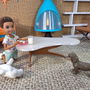 May include: A doll kneels near a mid-century modern style table with a glass of milk and a cookie. A brown dachshund dog stands nearby. A white stuffed dog lies on the floor. The scene is set in a mid-century modern style room.