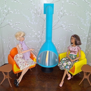 May include: Two dolls are seated in mid-century modern style chairs, one orange and one yellow, with small brown side tables. A tall, turquoise fireplace is between them. The dolls are wearing floral print dresses and accessories.