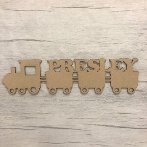 May include: Wooden train engine and carriages with the name "PRESLEY" cut out of the carriages. The train is brown and is on a light wood background.