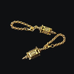 Puede incluir: Dos molinillos de oración de metal dorado con diseños intrincados están conectados por una cadena dorada. Los molinillos de oración tienen forma de pequeñas linternas con una punta puntiaguda en la parte inferior.