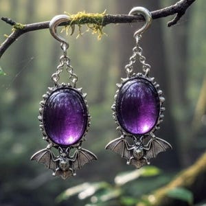 May include: A pair of silver-toned earrings with large, oval, purple gemstones. Each earring features a bat design below the gemstone. The earrings are suspended from a branch in a forest setting.