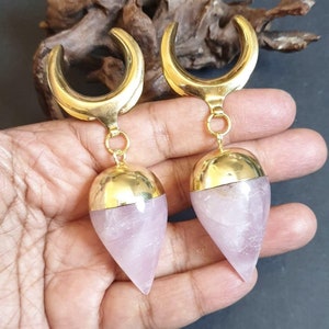 May include: A pair of gold-tone ear gauges with a crescent moon design and a pink quartz teardrop pendant.